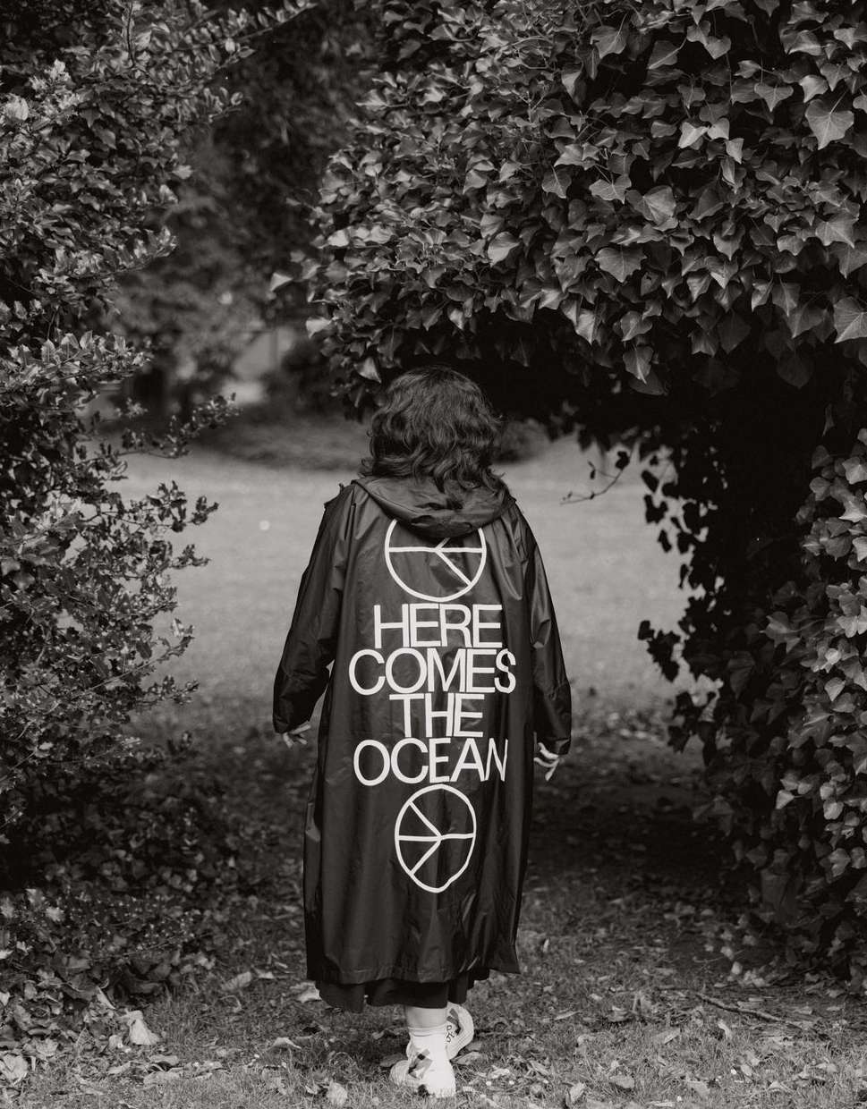 Woman wearing a black raincoat with 'Here Comes The Ocean' text and a peace symbol stands in an outdoor green, leafy environment.