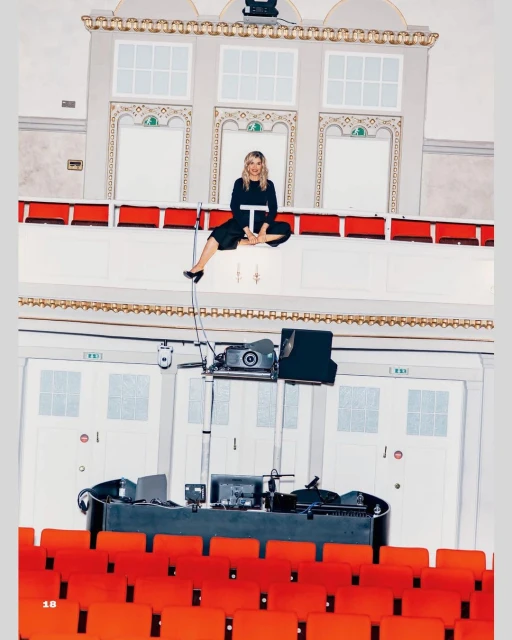 Blonde woman in casual clothing seated atop a ledge in an ornate theater or concert hall with rows of red seats below.