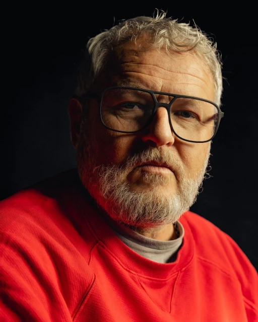 Portrait of a mature adult man with gray hair and beard, wearing glasses and a red sweatshirt against a dark background.