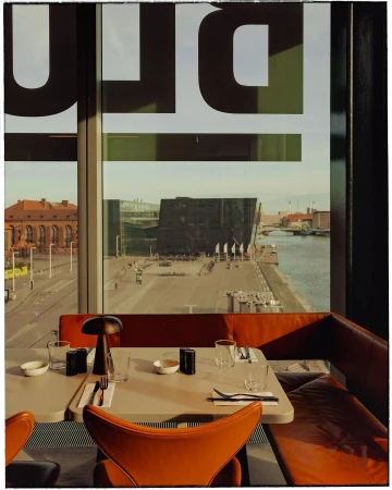 Interior shot of a restaurant with a harbor view featuring modern furniture, place settings, and large window graphics.
