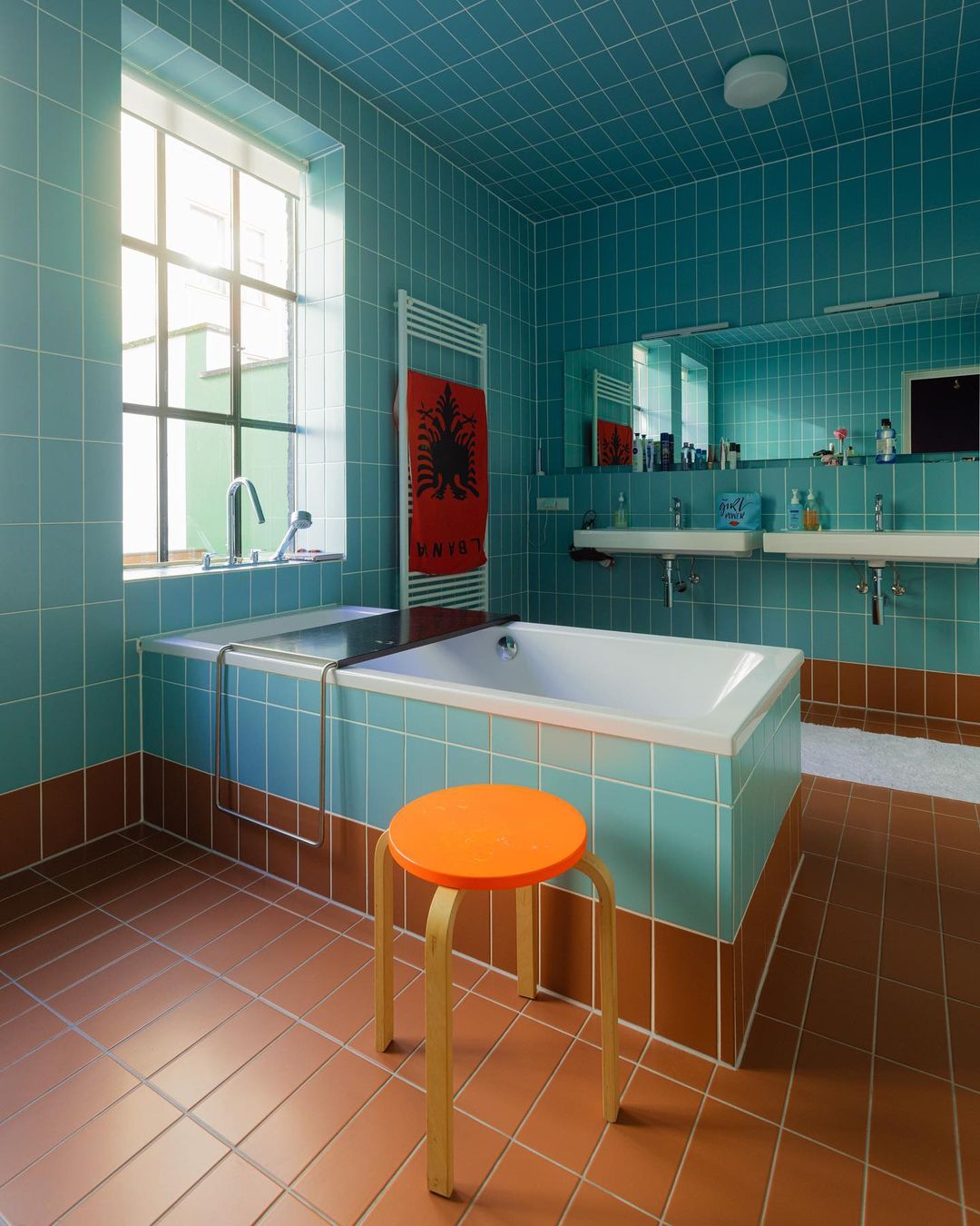 Retro bathroom interior featuring turquoise square tiling, terra cotta flooring, white bathtub, double sinks, and an orange stool.