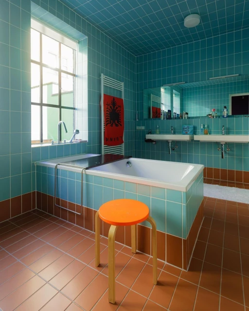 Retro bathroom interior featuring turquoise square tiling, terra cotta flooring, white bathtub, double sinks, and an orange stool.