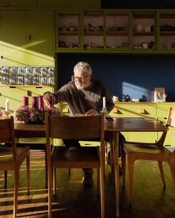 Senior adult man sitting at a table lighting purple advent candles, with green and blue kitchen cabinets in the background.