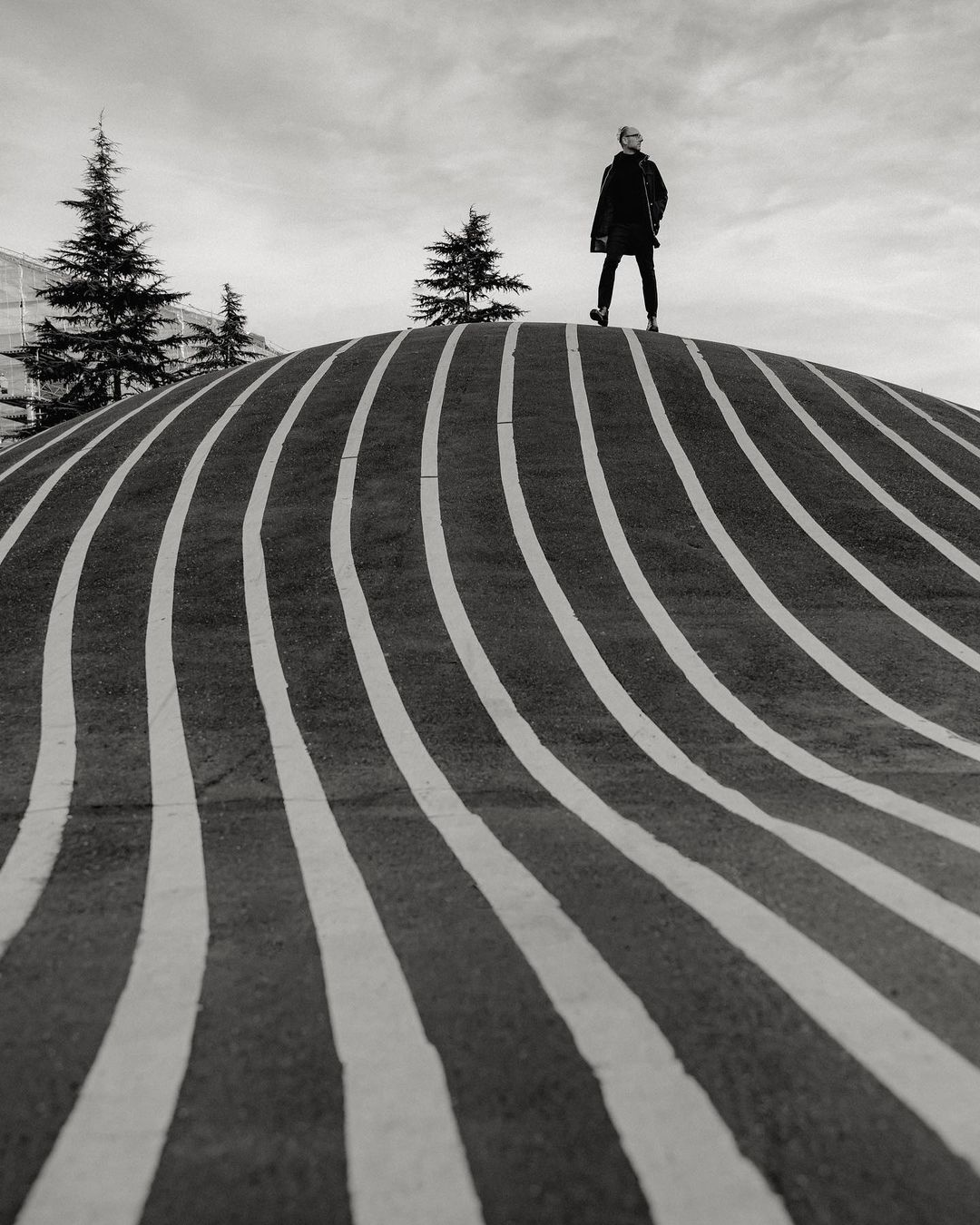Monochrome shot of a person standing on a striped, rounded structure with pine trees and cloudy sky in the background.