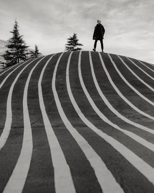 Monochrome shot of a person standing on a striped, rounded structure with pine trees and cloudy sky in the background.