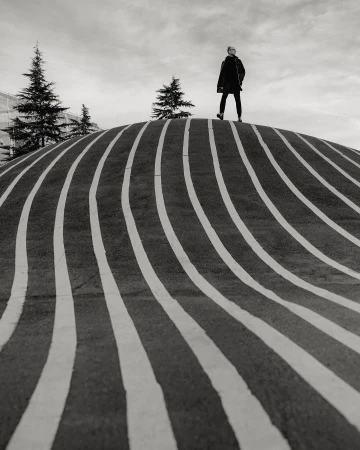 Monochrome shot of a person standing on a striped, rounded structure with pine trees and cloudy sky in the background.