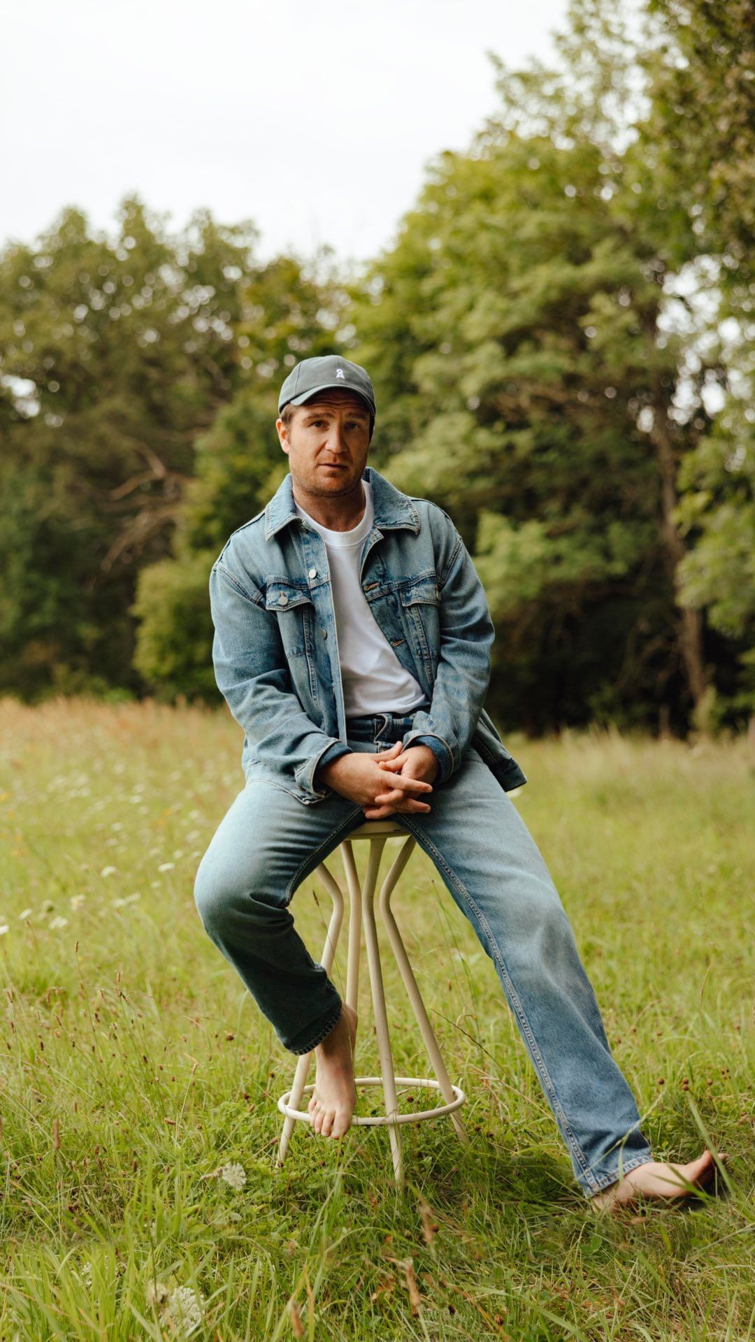 Man wearing denim jacket and cap seated on a stool in a grassy field by photographer Ramon Haindl