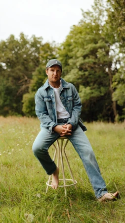 Man wearing denim jacket and cap seated on a stool in a grassy field by photographer Ramon Haindl
