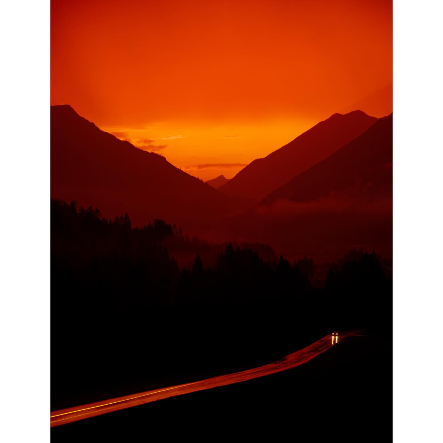 Scenic mountain range landscape at sunset with an orange sky, a road cutting through the dark forest, and car headlights.