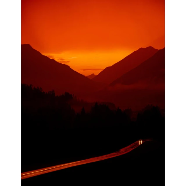 Scenic mountain range landscape at sunset with an orange sky, a road cutting through the dark forest, and car headlights.