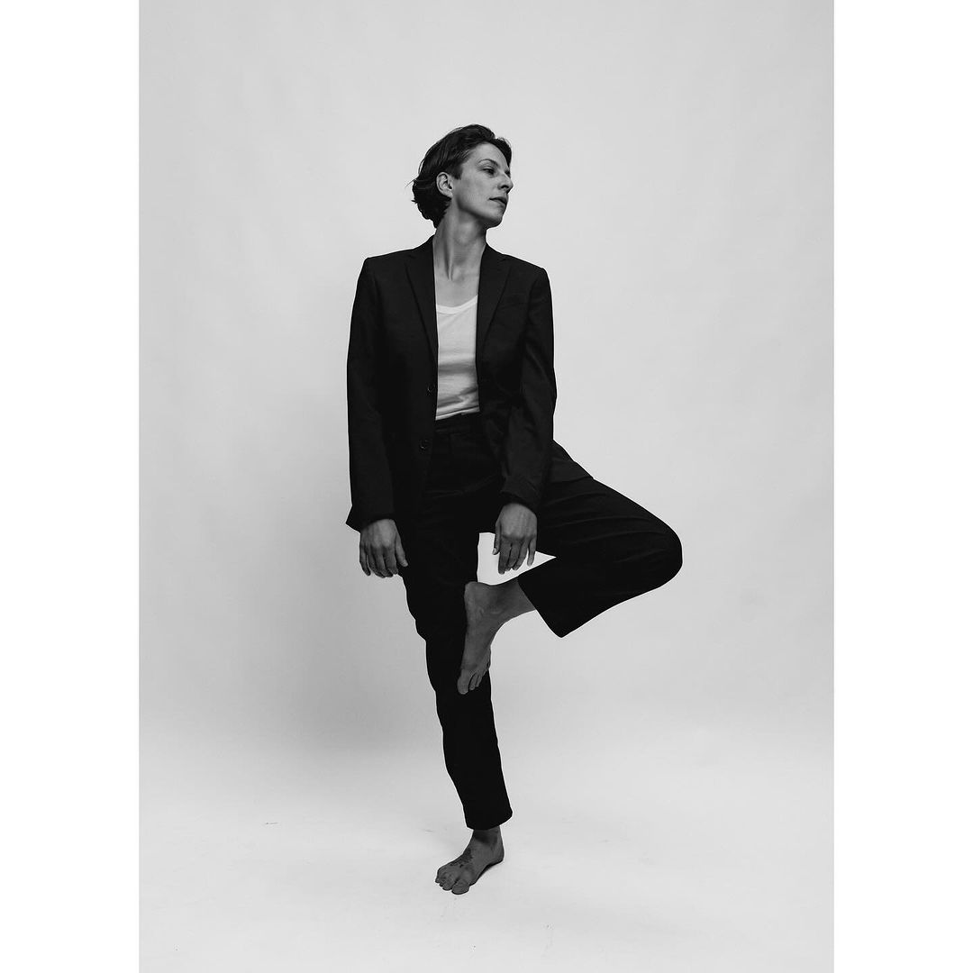 A woman in a dark suit is balancing on one leg against a neutral backdrop, creating a striking black and white portrait.