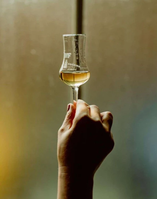 Woman's hand holding a clear glass of amber liquid with the brand "FOCKINK" visible against a blurred background.