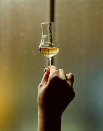 Woman's hand holding a clear glass of amber liquid with the brand "FOCKINK" visible against a blurred background.