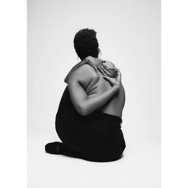 Back view of a nude African American woman seated on the floor with arms behind back in black and white photo studio portrait.
