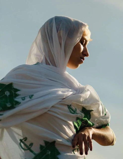 Woman in white headscarf with green design stands outdoors against a pale sky, creating a portrait with fashion and cultural elements.