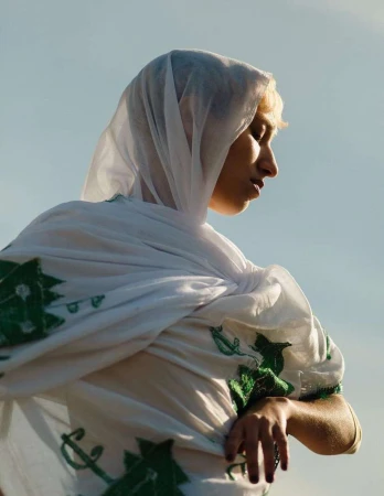 Woman in white headscarf with green design stands outdoors against a pale sky, creating a portrait with fashion and cultural elements.