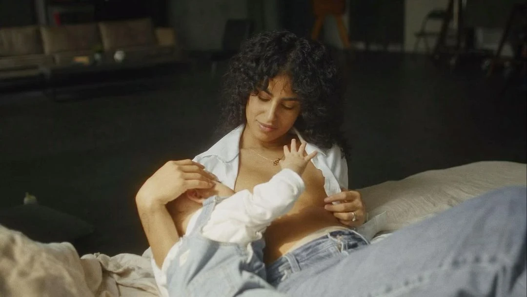 Mother with dark curly hair breastfeeding her baby indoors, wearing blue jeans and a white shirt, lying on a white blanket.