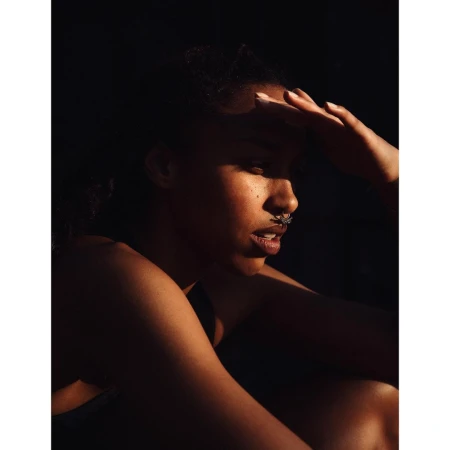 Portrait of a Black woman with a septum ring shielding her eyes in dramatic low-key lighting by Ramon Haindl.