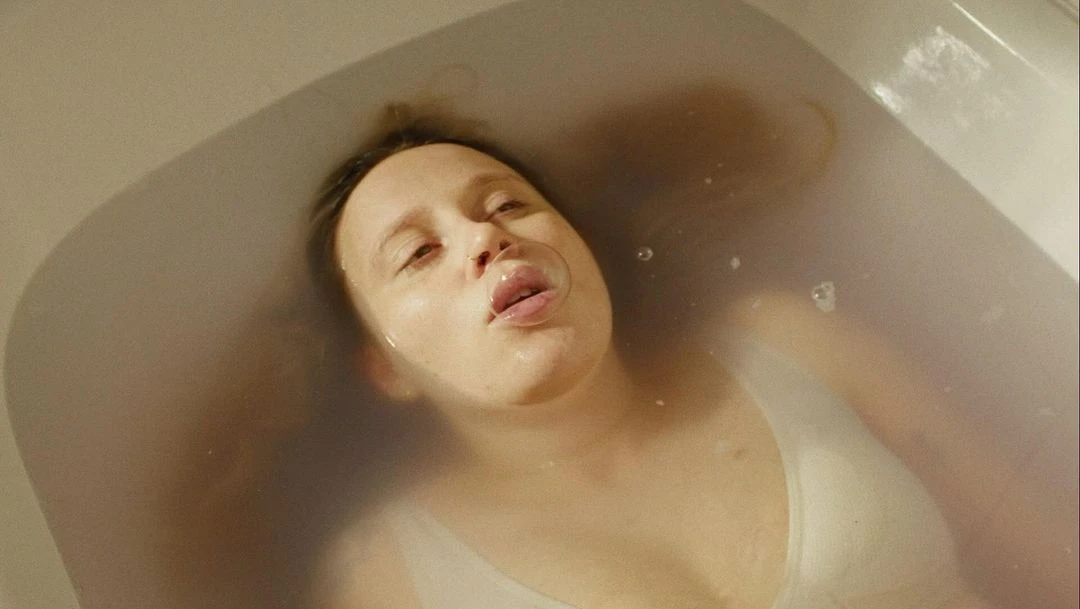Woman floats in bathtub with head partially submerged, looking upwards. Bubbles float around her face in a bathroom interior.