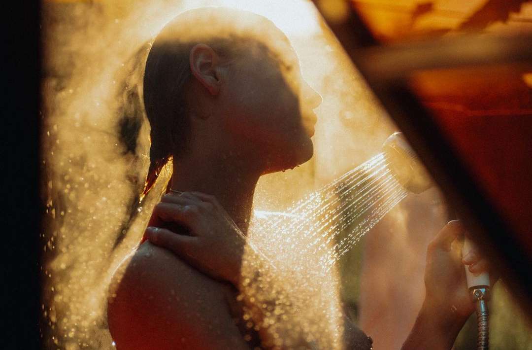 Woman with wet hair stands under shower head with water spraying, bathed in soft, golden light. Hand raised to shoulder.