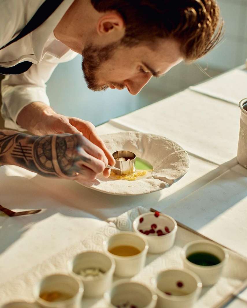 Close-up of a chef with arm tattoos garnishing a food plate, wearing a white shirt and black apron in an indoor food-prep setting.