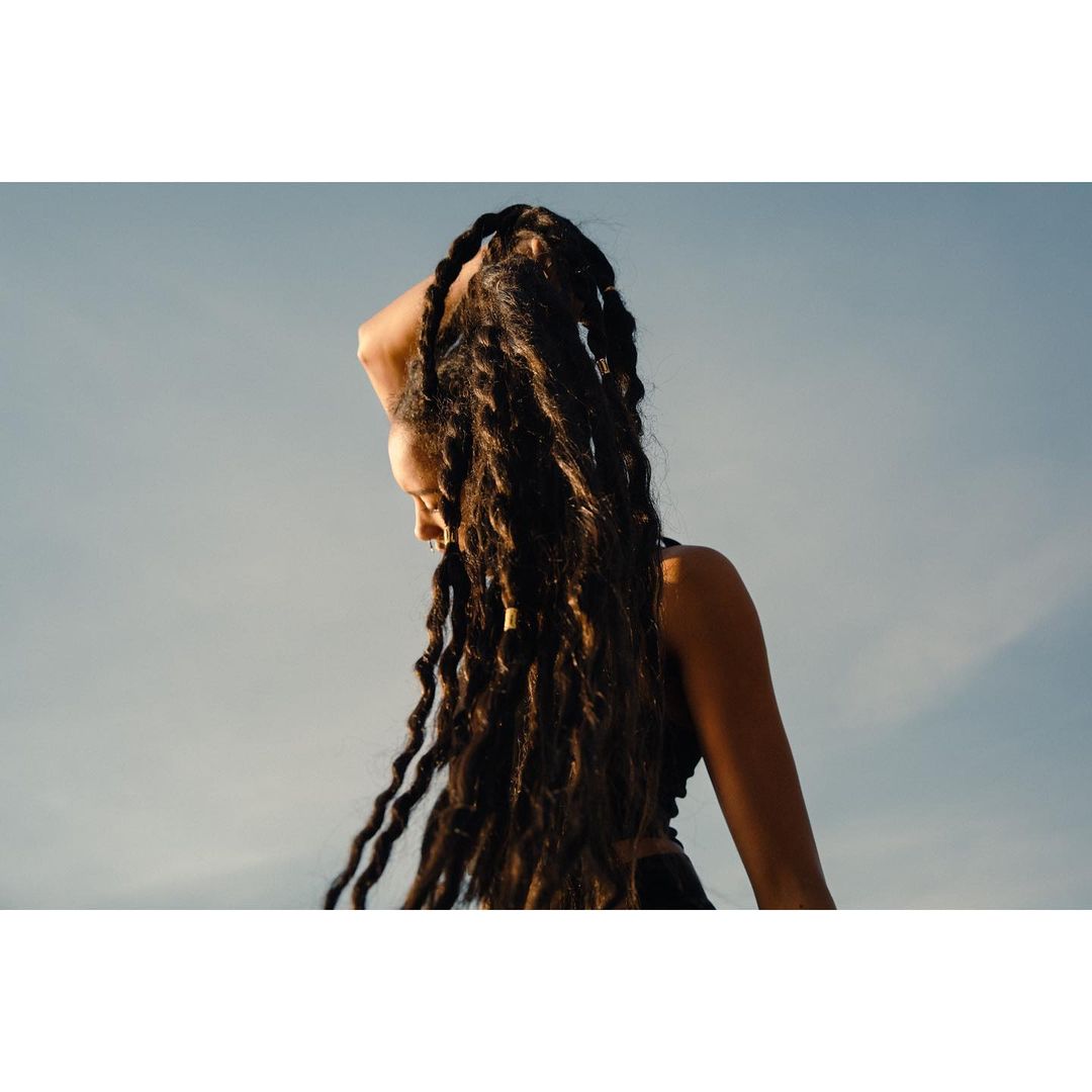 Portrait of an adult female with long dark dreadlocks, posing in an outdoor setting with soft lighting and a light blue sky.
