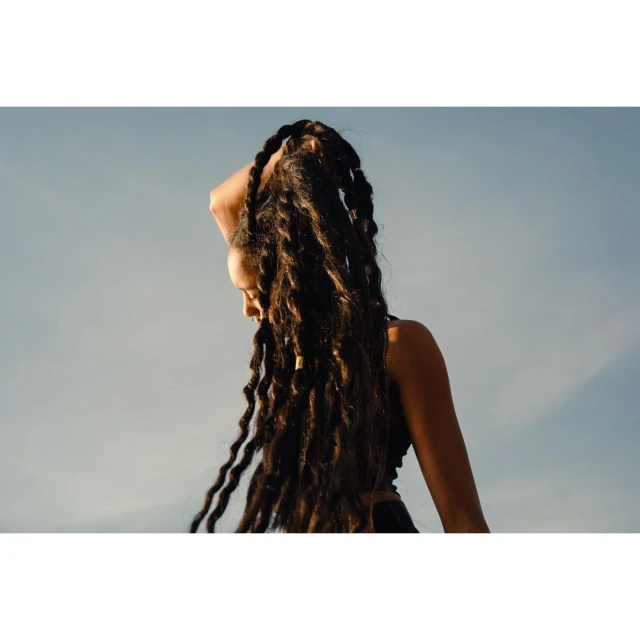 Portrait of an adult female with long dark dreadlocks, posing in an outdoor setting with soft lighting and a light blue sky.