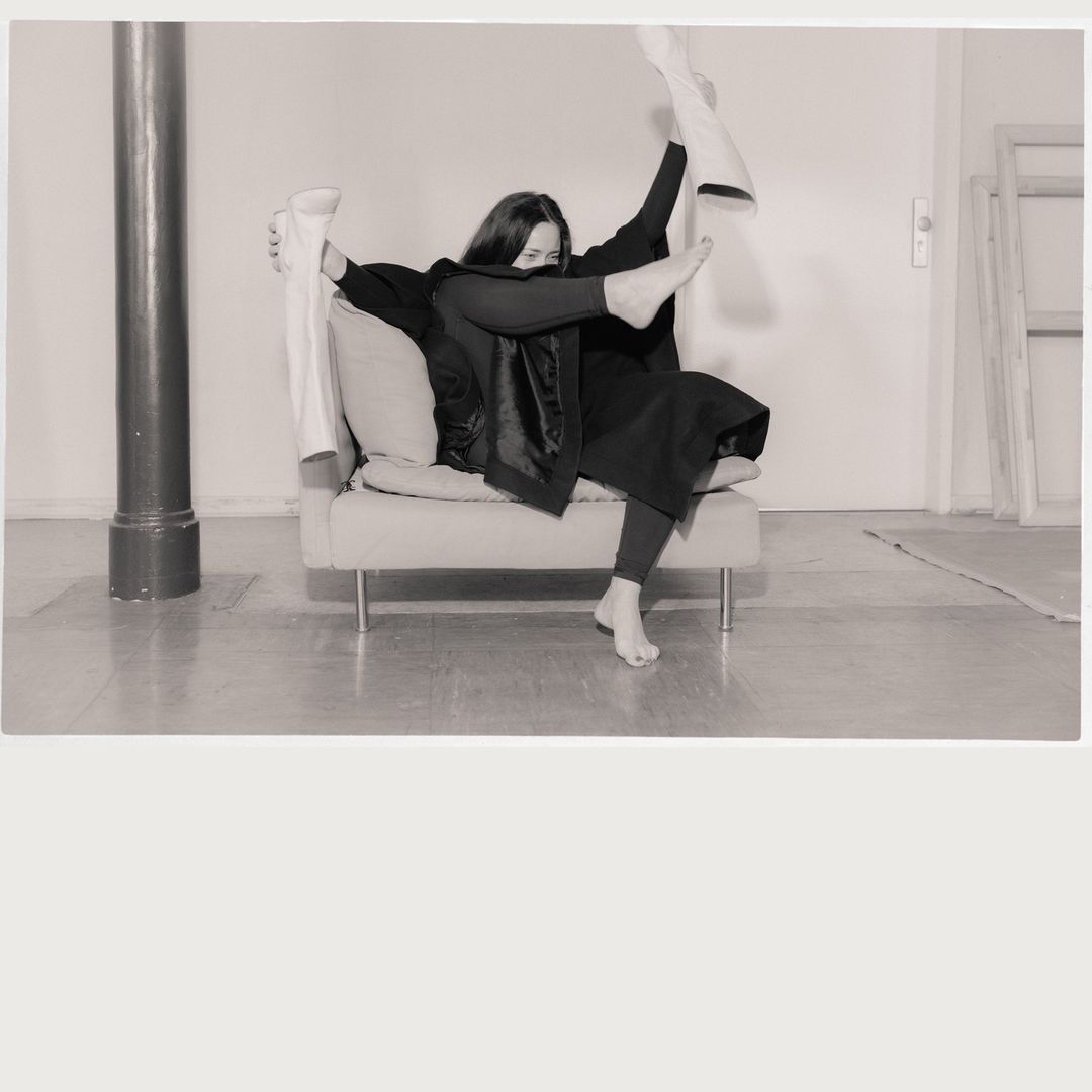 Monochrome indoor portrait of an adult female on a beige couch, playfully holding white boots, and wearing all black apparel.