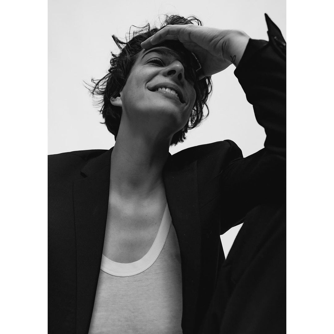 Low-angle portrait of a smiling adult with hand on head, wearing a blazer and tank top, set against a stark white background in black and white.