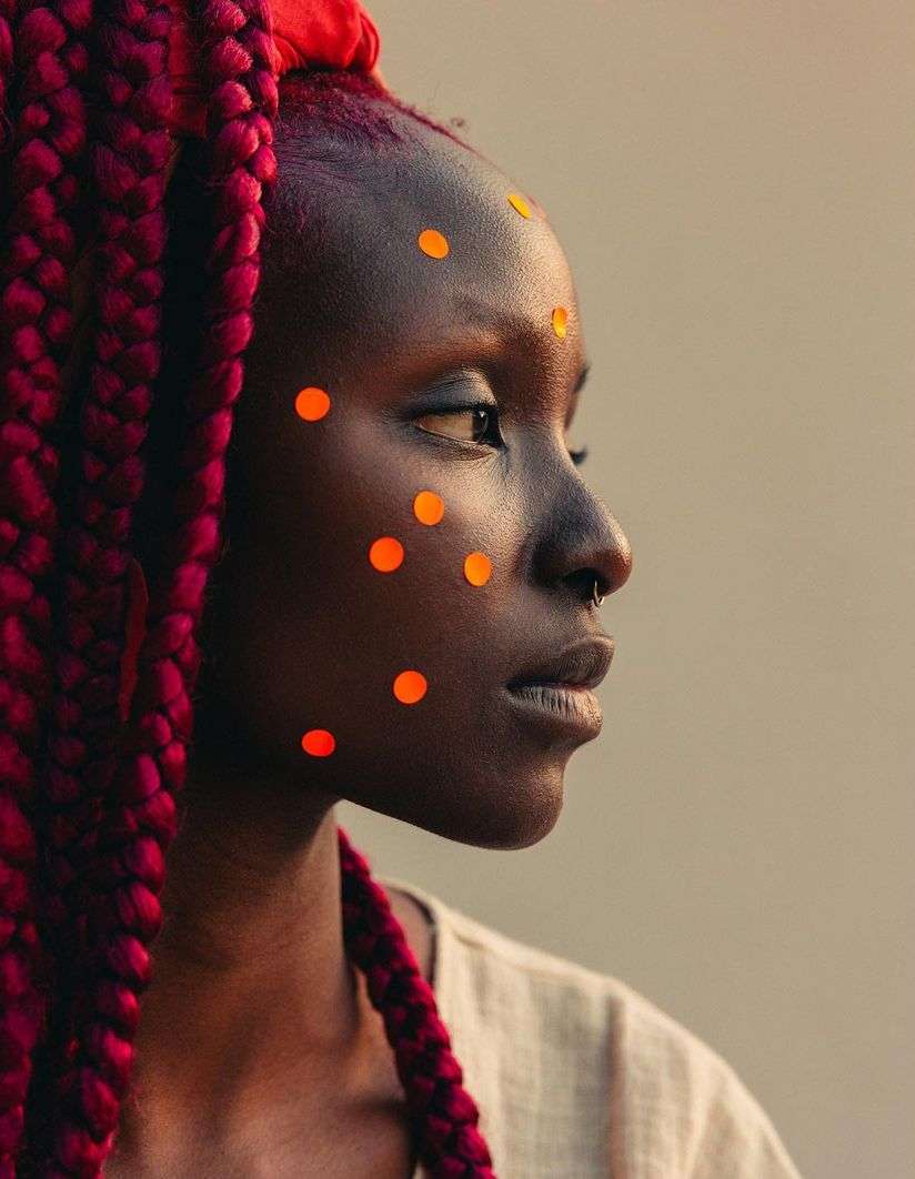 Close-up portrait of a dark-skinned woman with red braided hair, facing right, with orange dots on her face against a neutral backdrop.