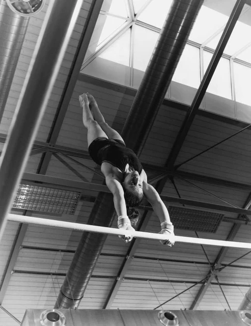A female gymnast executes a difficult move on parallel bars, shot from below in black and white, showcasing strength and skill indoors.