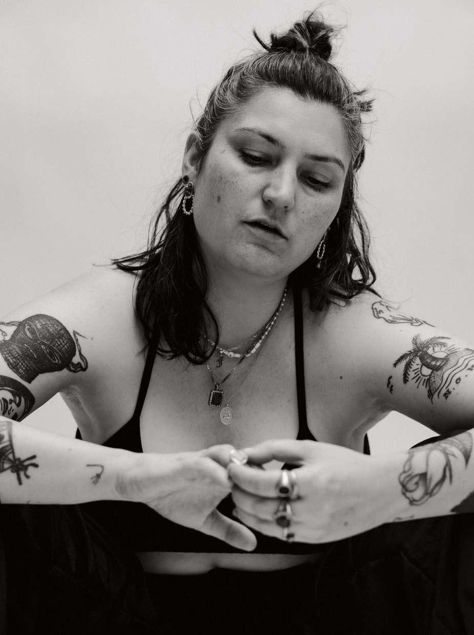 Black and white portrait of a young woman with multiple tattoos, silver necklaces and rings, and a dark top against a white backdrop.