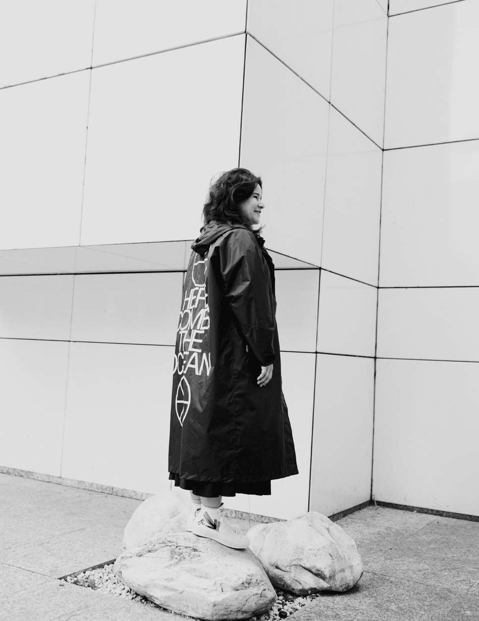 Adult female in dark raincoat stands on rocks in an urban environment. The text on the back of the raincoat reads 'Here come the ocean'.