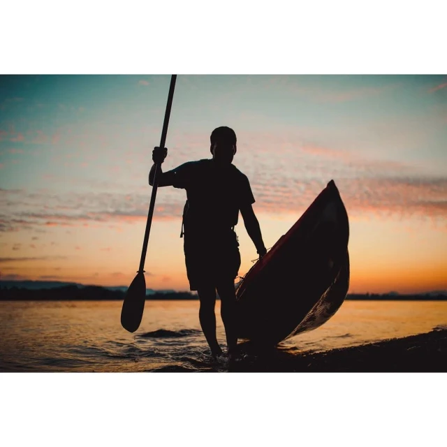 Silhouette of adult carrying canoe and paddle walking on shoreline at sunset, creating dramatic, colorful, outdoor watersport image.