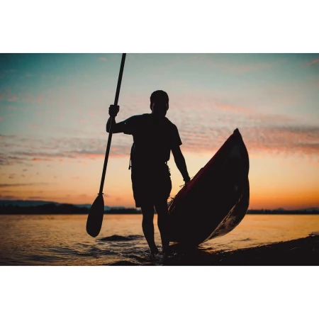 Silhouette of adult carrying canoe and paddle walking on shoreline at sunset, creating dramatic, colorful, outdoor watersport image.