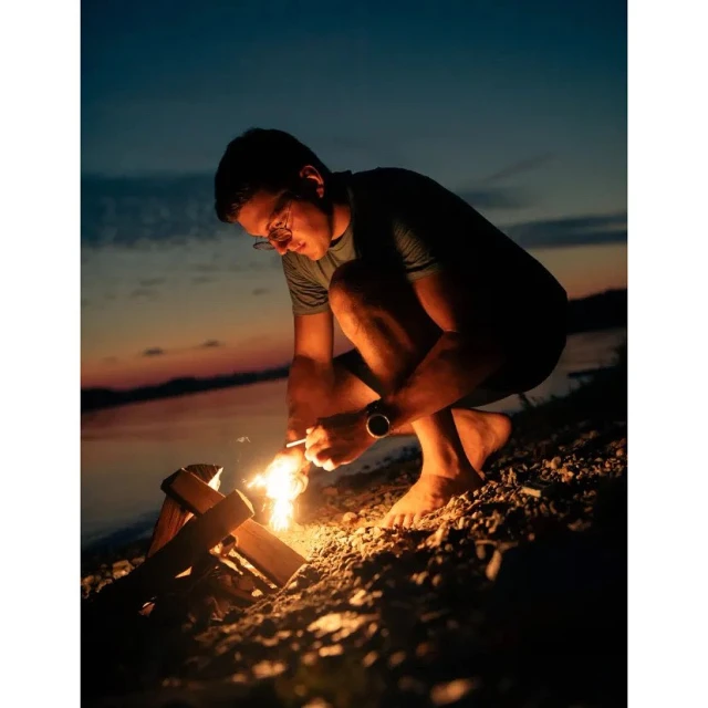 Man squats barefoot on a beach at dusk, lighting a small campfire made of driftwood with a match, with water in background.