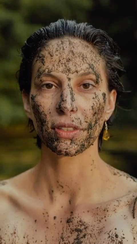 Close-up studio portrait of a woman with dark hair and an ambiguous expression, her face and upper body covered in wet soil or mud.