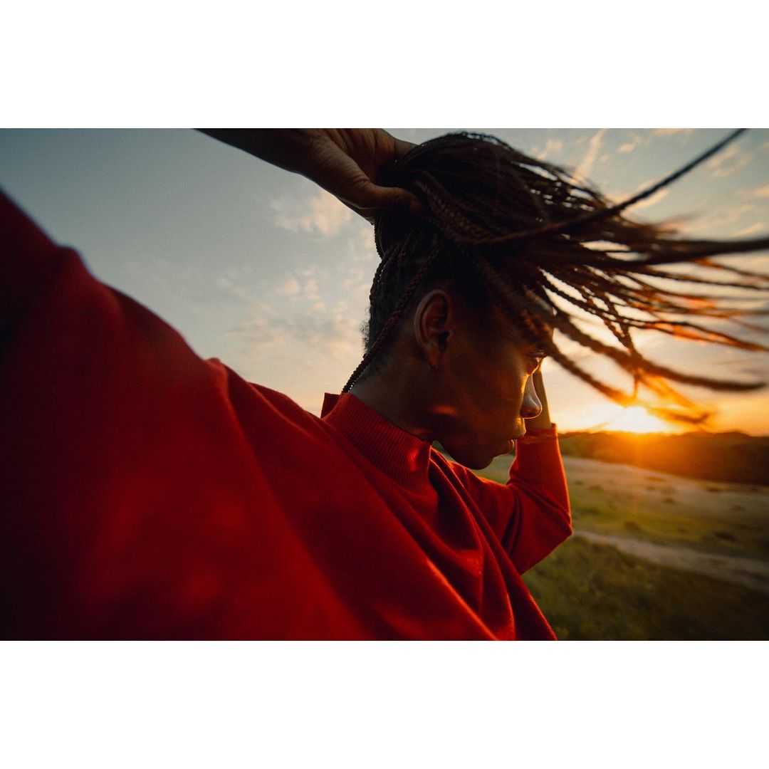 Woman in a red sweatshirt standing outside with her hair blowing in the wind at sunset in a field. Warm tones and natural lighting.