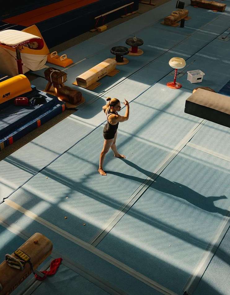Female gymnast performs a routine in a sunlit gymnasium with blue mats and equipment, casting long shadows in an overhead shot.