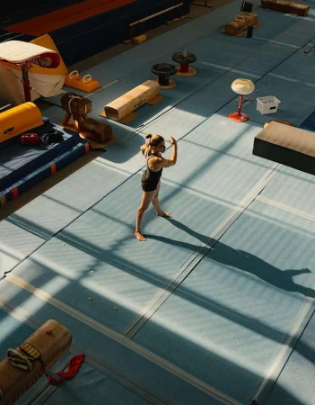 Female gymnast performs a routine in a sunlit gymnasium with blue mats and equipment, casting long shadows in an overhead shot.