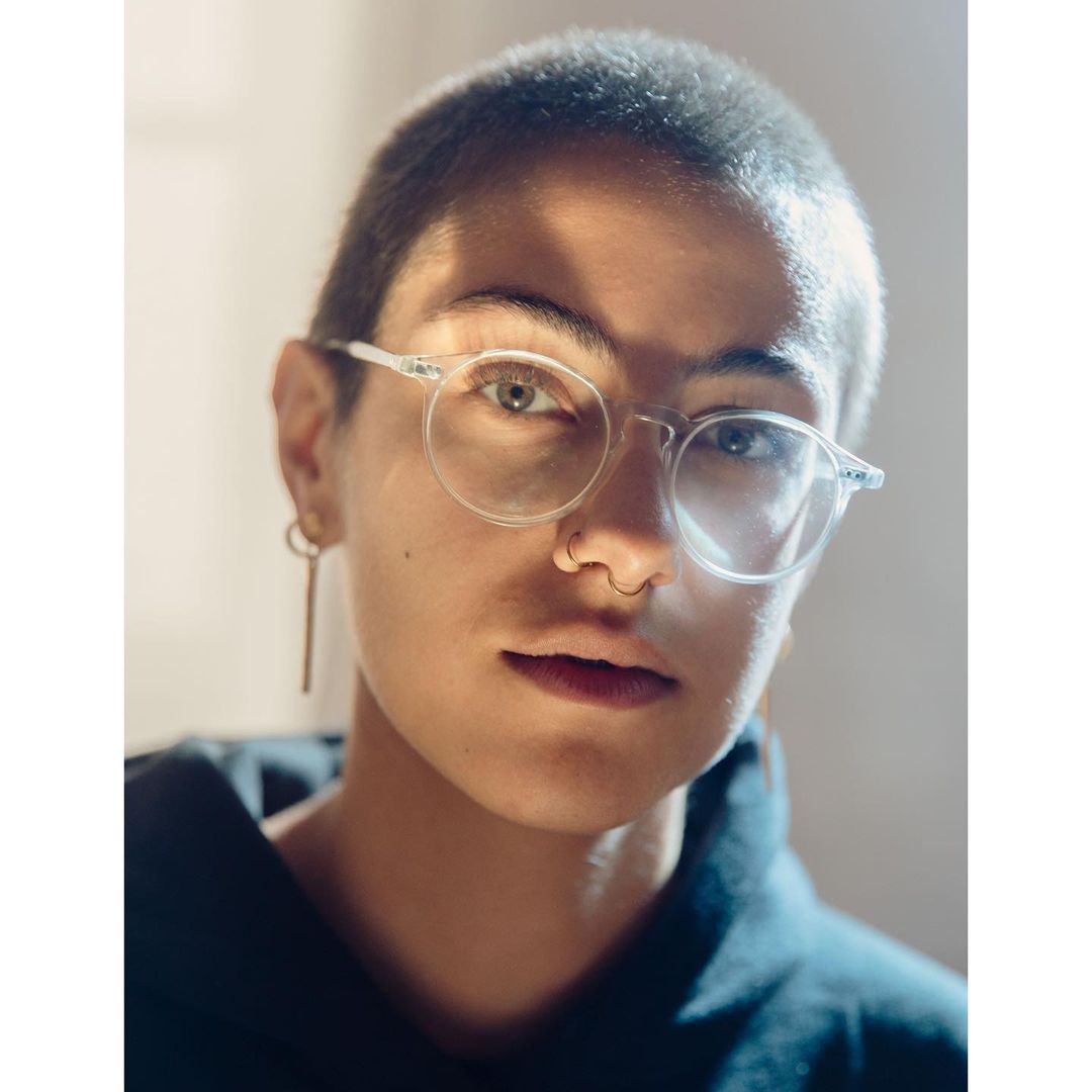 Portrait of a young adult female with short hair, wearing clear glasses and a dark top, with warm light illuminating her face.