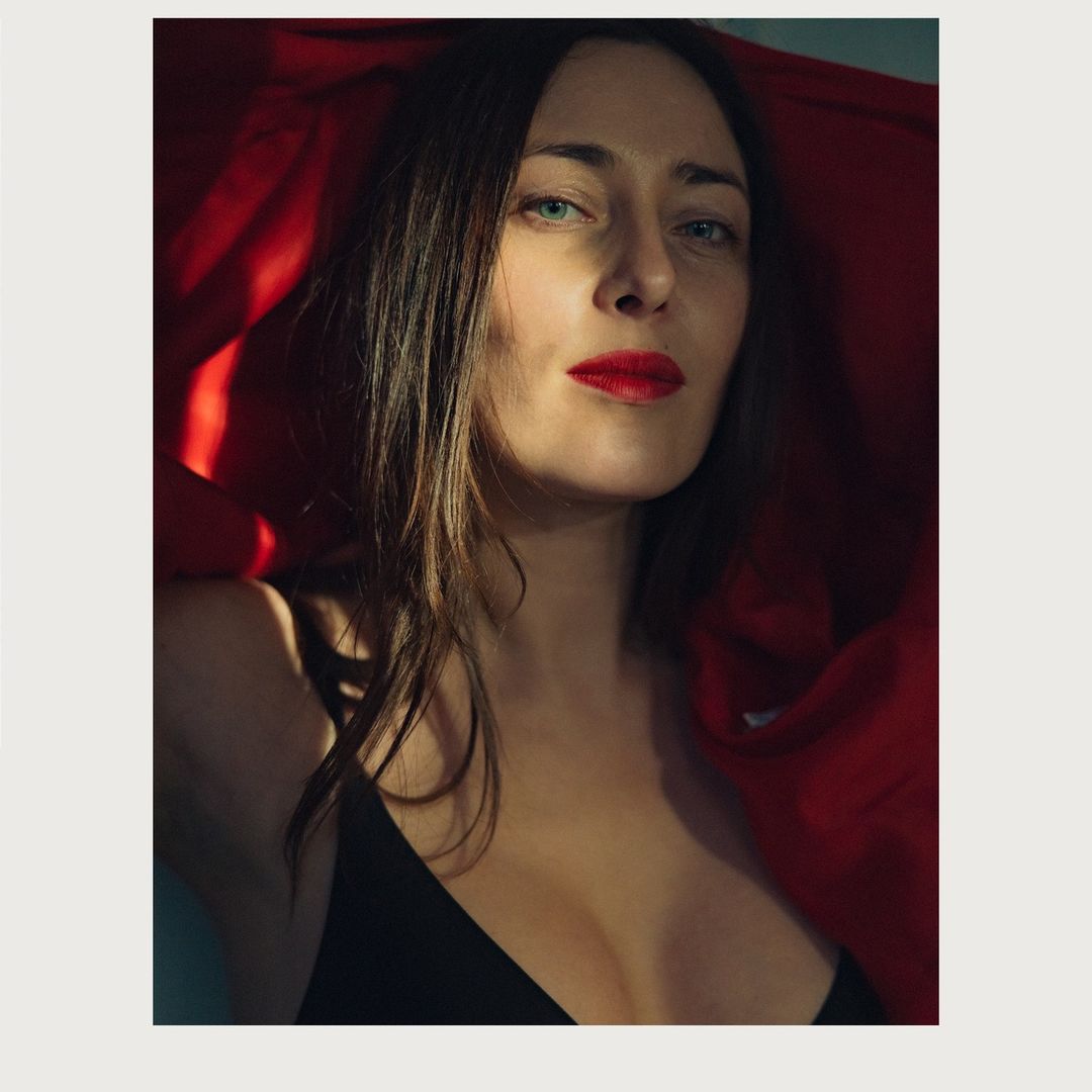 Headshot of a fair-skinned woman with long brunette hair and bright red lipstick, wearing a black top and red jacket in a studio.