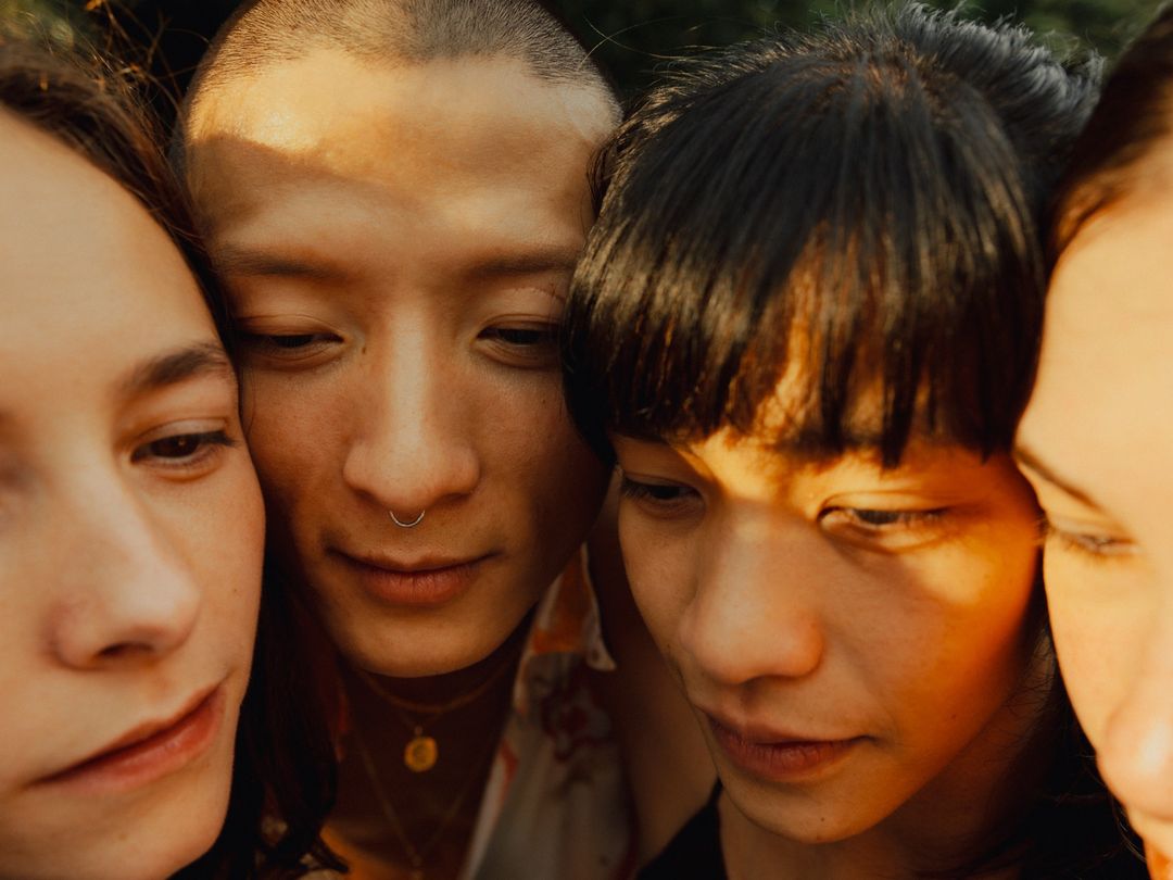 Close-up of four young adults, diverse ethnicities, gathered closely together, heads touching. Warm light, shallow depth of field.