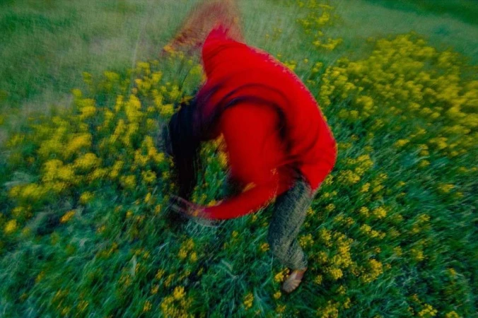 Blurred image of a person in a red hooded jacket crouching in a field of green grass and yellow flowers, captured with motion blur.