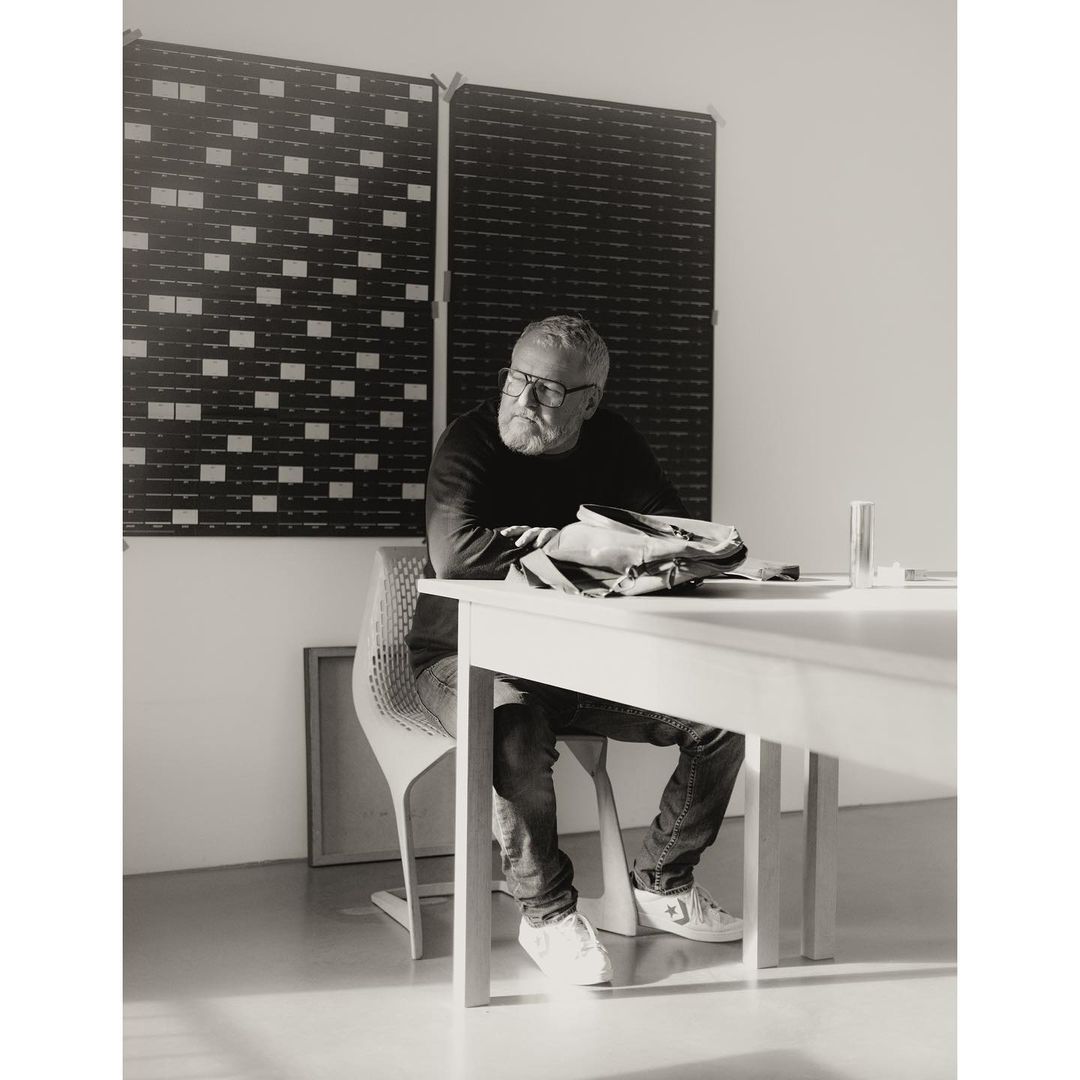 Monochrome portrait of an adult male seated at a white table with a black and white textured backdrop, wearing glasses and casual clothing.
