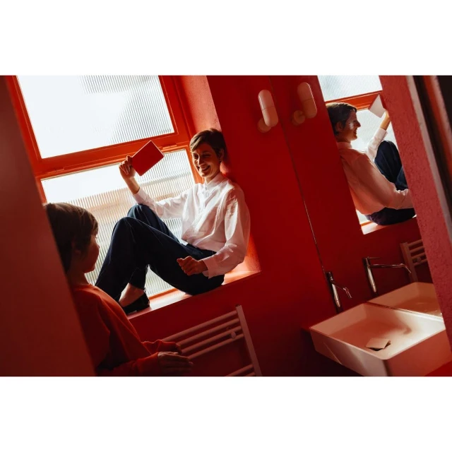 A young woman sits on a window ledge holding a book, with a child and bathroom sink visible in the foreground of the red room.