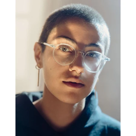 Portrait of a person with a buzzcut and clear round glasses wearing a dark hoodie, photographed by Ramon Haindl