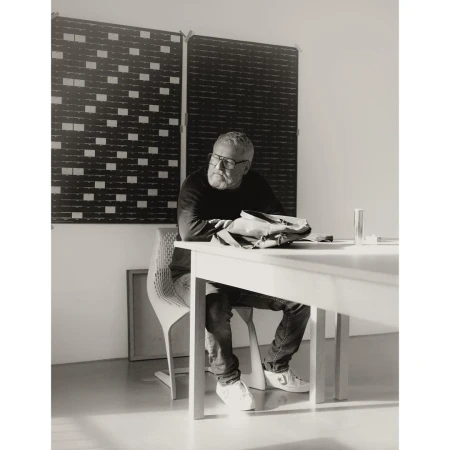 Man wearing glasses seated at a white table in a bright room with dark patterned acoustic panels on the wall.