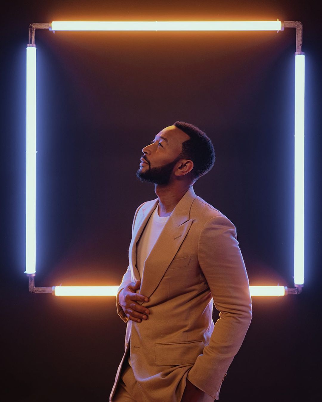 Portrait of a Black adult male in a tan suit looking upward, standing against a dark background with illuminated square lighting.