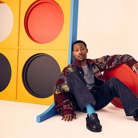 A Black man seated on the floor wearing a patterned burgundy jacket, grey jumper, wide jeans, and bright blue socks.